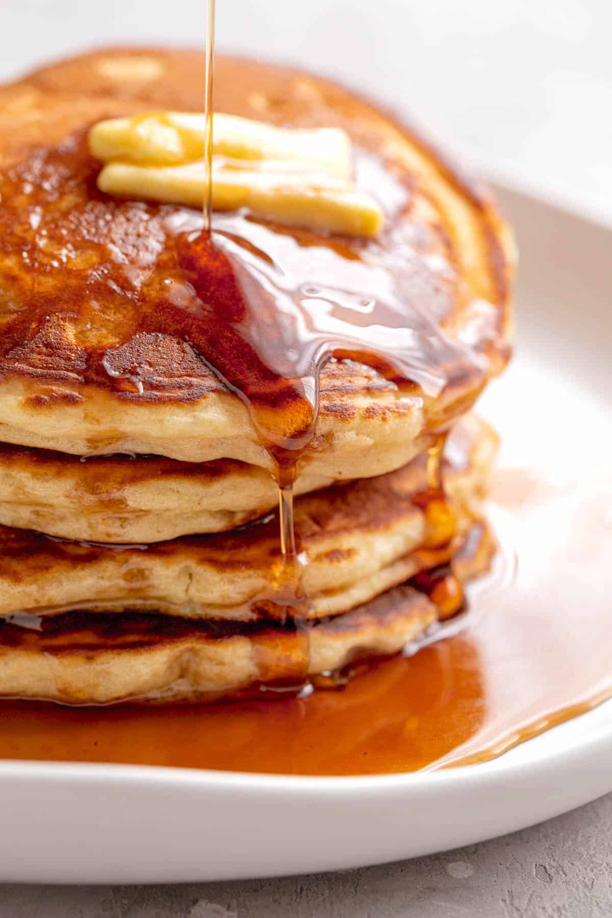 A stack of Fluffy Pancakes on a white plate served with butter and drizzled with maple syrup. | craveitall.com